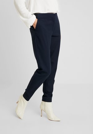 Person iført marineblå smalle bukser med hænderne i lommerne og hvide ankelstøvler med hæl, stående mod en ensartet lys baggrund.