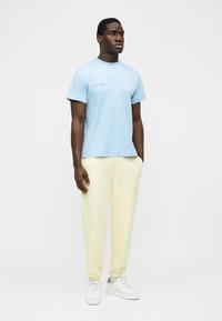 Młody mężczyzna stoi, ubrany w jasnoniebieską koszulkę, kremowe spodnie dresowe i białe trampki na jednolitym białym tle.