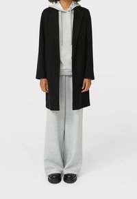 Manteau long noir sur un sweat à capuche gris et un pantalon de survêtement large. Présente une coupe décontractée et un tissu texturé. Des chaussures noires épaisses complètent la tenue.