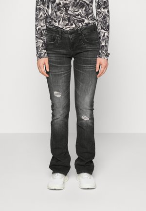 Personne portant un jean noir délavé et usé, des baskets blanches et un haut à manches longues avec un motif abstrait noir et blanc sur un fond uni.