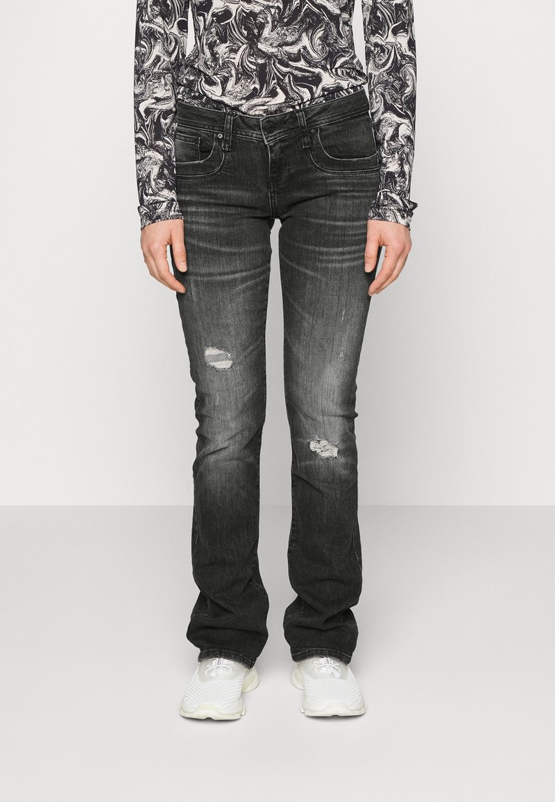 Personne portant un jean noir délavé et usé, des baskets blanches et un haut à manches longues avec un motif abstrait noir et blanc sur un fond uni.