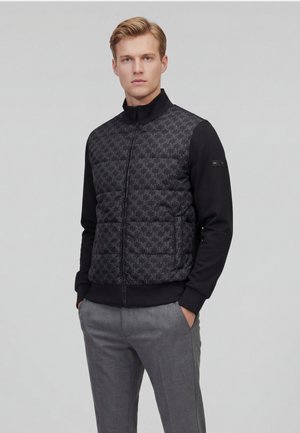 Mand iført sort lynlåstrøje med mønstret forside og grå bukser, stående med hænderne i lommerne mod en ensfarvet baggrund.