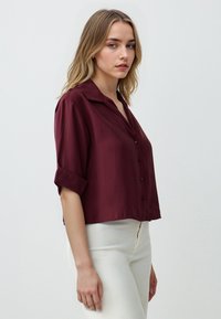 Burgunderfarbige Bluse mit kurzen Ärmeln, Kragen, Knopfleiste und umgeschlagenen Ärmeln; weicher Stoff und lockere Passform, kombiniert mit weißen Hosen.