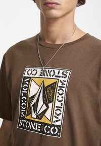 Brun T-shirt med et stort grafisk design i sort, hvid og gul med tekst. Korte ærmer og rund halsudskæring.