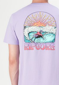 Fialová bavlnená tričko s grafickým motívom surfovania, znázorňujúce vlny a slnko, s textom "RIPCURL" v tučnom, farebnom písme na spodnej časti.