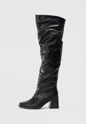 Black knee-high boots made of shiny leather with a slouchy design, pointed toe, and a chunky block heel. Smooth texture with subtle shine.