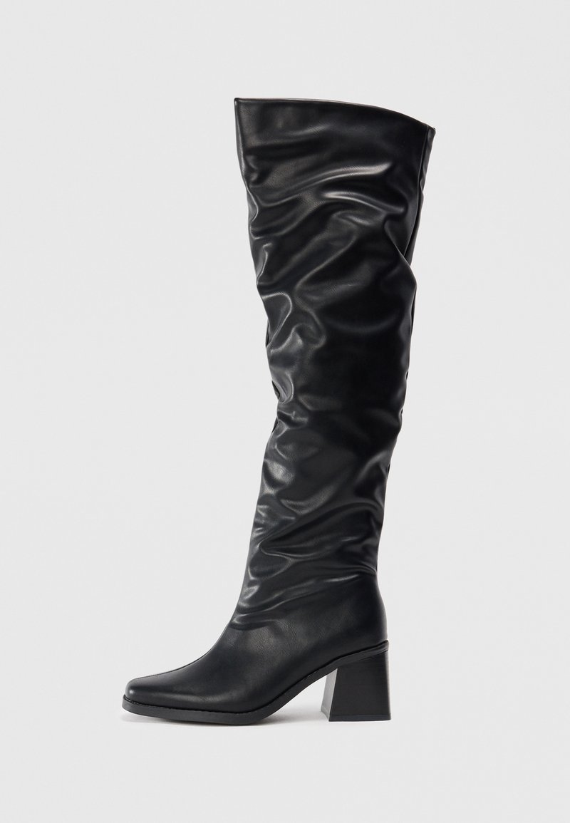 Bottes noires montantes en cuir brillant avec un design froncé, bout pointu et talon block chunky. Texture lisse avec un léger éclat.