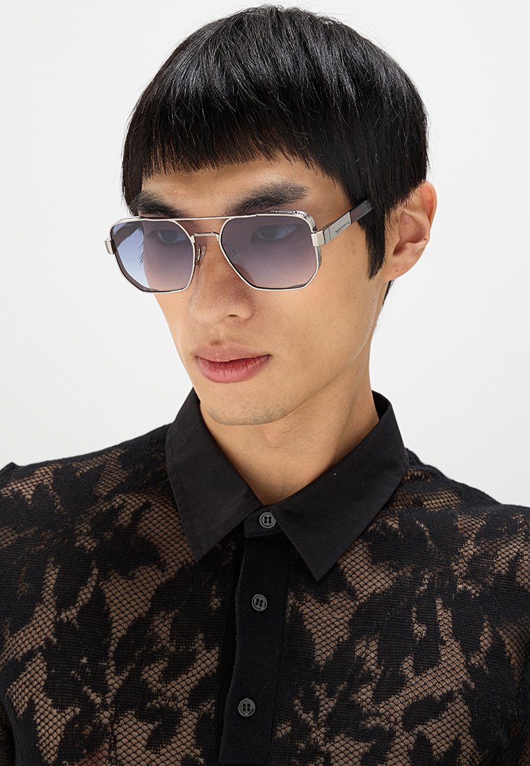 Solglasögon med en silverfärgad ram, fyrkantig form och graderade lila linser. Modellen bär en svart, genomskinlig spetsblus med krage.