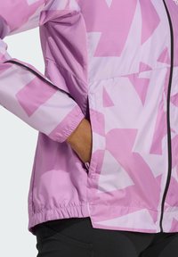 Lätt rosa jacka med djärva geometriska mönster, har dragkedjeförslutning, meshpaneler och elastiska ärmslut. Sidoficka synlig.