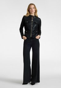 Giacca in pelle nera con dettagli dorati e tasche a pattina frontali, abbinata a jeans a gamba larga scuri. Il modello sta in piedi con le mani nelle tasche.