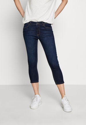 Osoba ubrana w ciemnoniebieskie, krótkie rurki, białe trampki i luźną białą koszulkę, stojąca na jasnym, jednolitym tle.