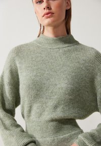 Pull en tricot de couleur vert clair avec un col haut à côtes. Il présente un motif texturé et des manches amples, fabriqué à partir d'un matériau doux.