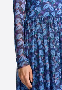 Vestido floral con largas mangas transparentes y una falda plisada. Predominantemente azul con patrones florales morados y verdes sobre un fondo oscuro.