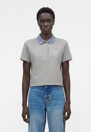 Femme portant un polo gris à manches courtes avec un col bleu et un jean bleu sur un fond blanc uni.