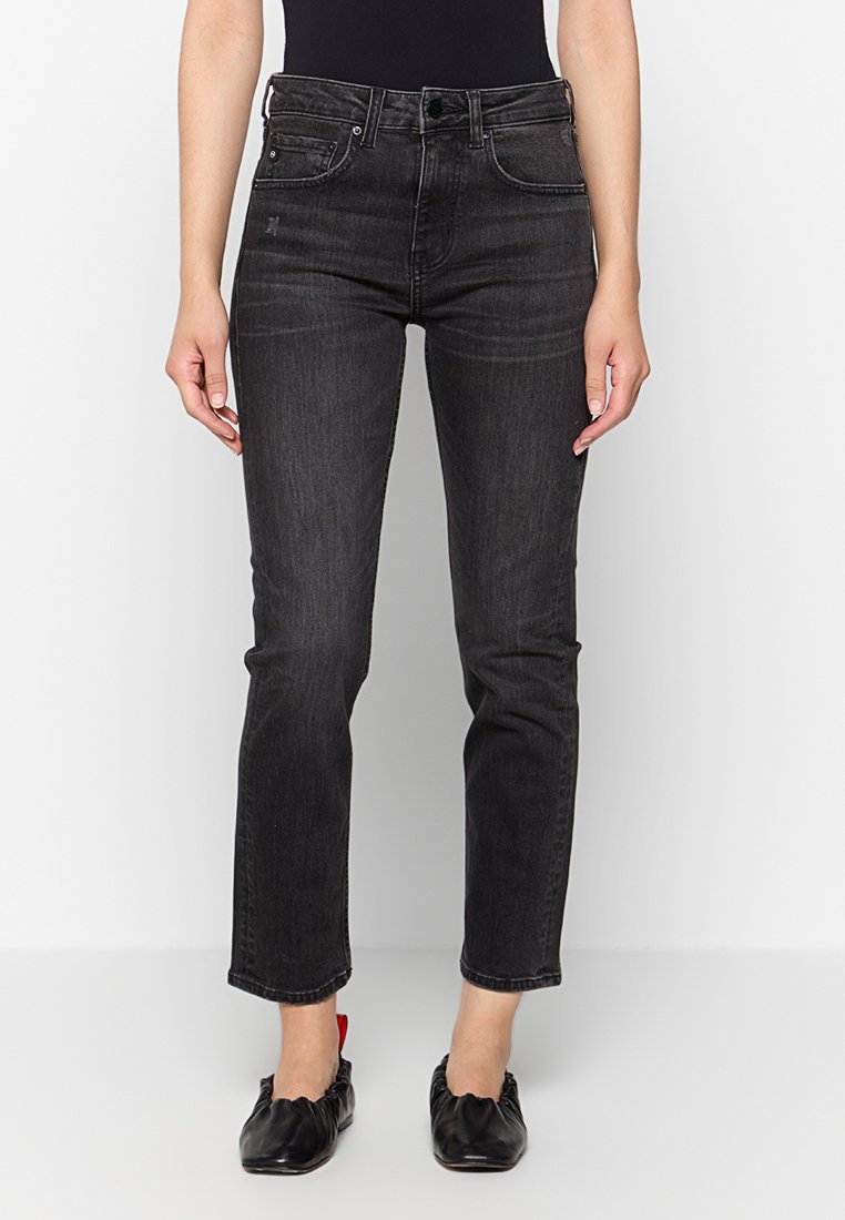 Kobieta w czarnych, dopasowanych jeansach z wysokim stanem, sięgających powyżej kostek, w połączeniu z czarnymi skórzanymi baletkami z czerwonym detalem na pięcie.