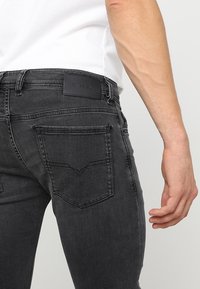 Jeans de mezclilla negra con un corte ajustado, que presentan un parche de cuero etiquetado como "DIESEL", dos bolsillos traseros y detalles de costura sutiles.