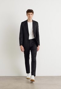 Črn suit z natančnim krojem, nosi se čez belo majico, v kombinaciji s slim črnimi hlačami in belimi supergami. Minimalistična zasnova in gladka tekstura.