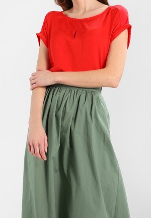 Vrouw draagt een rood kortarmig blouse met opgerolde manchetten en een olijfgroene, hooggetailleerde rok met plooien, armen gekruist ter hoogte van de taille.