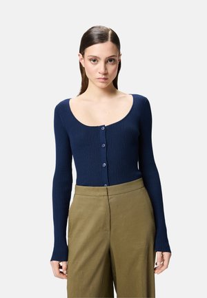 Cardigan lungo a coste di colore blu navy con scollatura a girocollo e cinque bottoni, abbinato a pantaloni oliva a vita alta.