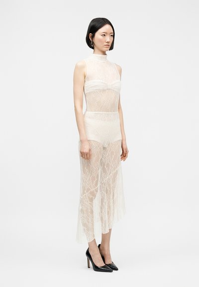 Γυναίκα που φοράει αμάνικο, διάφανο λευκό δαντελωτό φόρεμα με ψηλό λαιμό, λευκά εσώρουχα και μαύρα ψηλοτάκουνα παπούτσια, στέκεται σε απλό φόντο.