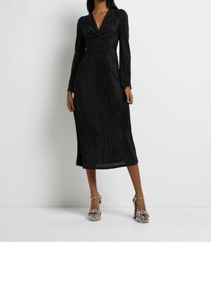 Femme portant une longue robe noire plissée à manches longues, assortie à des sandales à talons hauts imprimé zèbre noir et blanc.