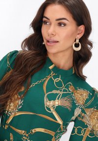 Zöld ruha aranylánc és virágmintákkal. Magas nyak, pufi ujjak és részletes díszítések. A modell nagy, texturált karika fülbevalókat visel.
