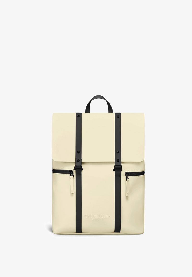 Sac à dos couleur crème avec des sangles noires, deux poches avant zippées, une poignée supérieure et une fermeture à rabat, embossé du texte "Gaston Luga Stockholm".