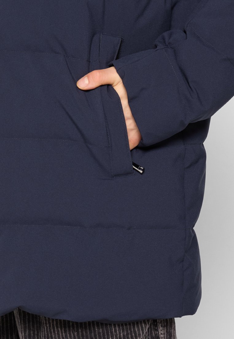 Hand in der Tasche einer marineblauen, gepolsterten Jacke mit sichtbar herausziehbarem Reißverschluss am Taschenrand.