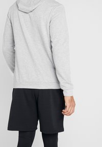Homme portant un sweat à capuche gris clair, un short noir et des leggings noirs, vu de dos avec la main droite détendue sur le côté.