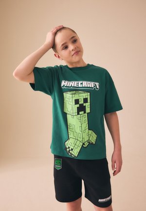 Dreng med kort hår iført en grøn Minecraft T-shirt og sorte shorts, stående med den ene hånd på hovedet mod en ensfarvet baggrund.