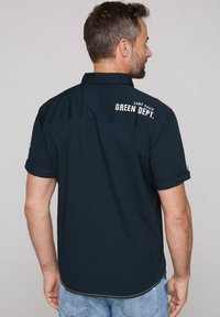 Navy short-sleeved shirt made of smooth fabric, featuring a curved hem, embroidered “CAMP DAVID GREEN DEPT.” text, and rolled cuff details.