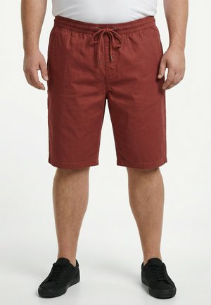 Mann trägt rostfarbene Shorts mit Kordelzug und schwarze Turnschuhe und steht vor einem schlichten Hintergrund.