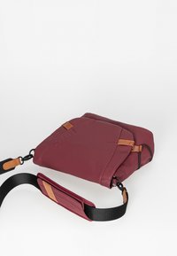 Bordeaux farvet crossbody taske med en glat tekstur, dekoreret med brune læderdetaljer, justerbar rem og et kompakt, rektangulært design.