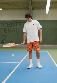 T-shirt bianco, pantaloni corti a quadri arancioni, con una racchetta da tennis in mano, in piedi su un campo blu, con uno sfondo verde e una pallina da tennis nelle vicinanze.