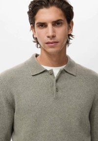 Maglione polo grigio chiaro in lana con colletto, due bottoni e una texture sottile. Indossato sopra una maglietta bianca. Vistao ravvicinato del colletto.