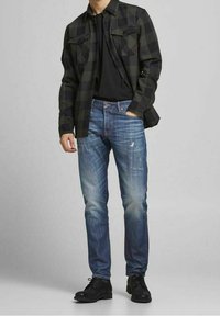 Camisa de franela de cuadros en negro y verde, combinada con una camiseta negra de cuello redondo y jeans desgastados de color azul claro. Zapatos negros completan el atuendo.