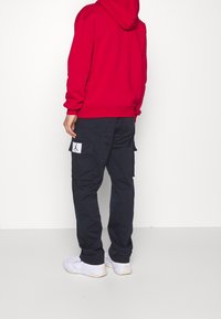 Rode capuchonsweatshirt met geribbelde manchetten, gecombineerd met marineblauwe cargobroek met zijzakken en een klein logoplaatje, gedragen met witte sneakers.