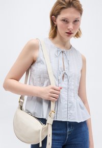 Γυναίκα που φορά αμάνικο λευκό μπλουζάκι και μπλε τζιν, κρατώντας μια κρεμ δερμάτινη τσάντα ώμου με αγκράφα σε χρυσή απόχρωση.