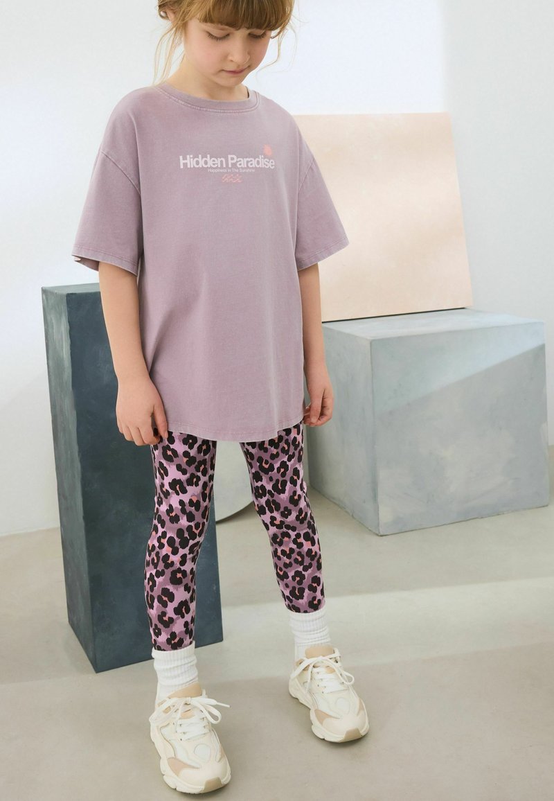 Lila oversized t-shirt med texten "Hidden Paradise", tillsammans med rosa leopardmönstrade leggings och vita sneakers med texturerade sulor.