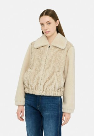 Giacca in pelliccia sintetica beige con texture a maglia cable-knit, colletto e chiusura con zip. Presenta un orlo elasticizzato e tasche laterali. Abbinata a jeans blu.