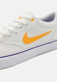 Bílé plátěné tenisky s oranžovým logem Nike, šněrovací design, polstrovaný límec, fialový pruh na podrážce a texturovaná gumová podrážka.