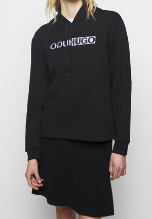 Osoba nosząca czarną bluzę z kapturem z odwróconym fioletowym napisem "HUGO" i czarną teksturowaną spódnicą na jednolitym jasnym tle.
