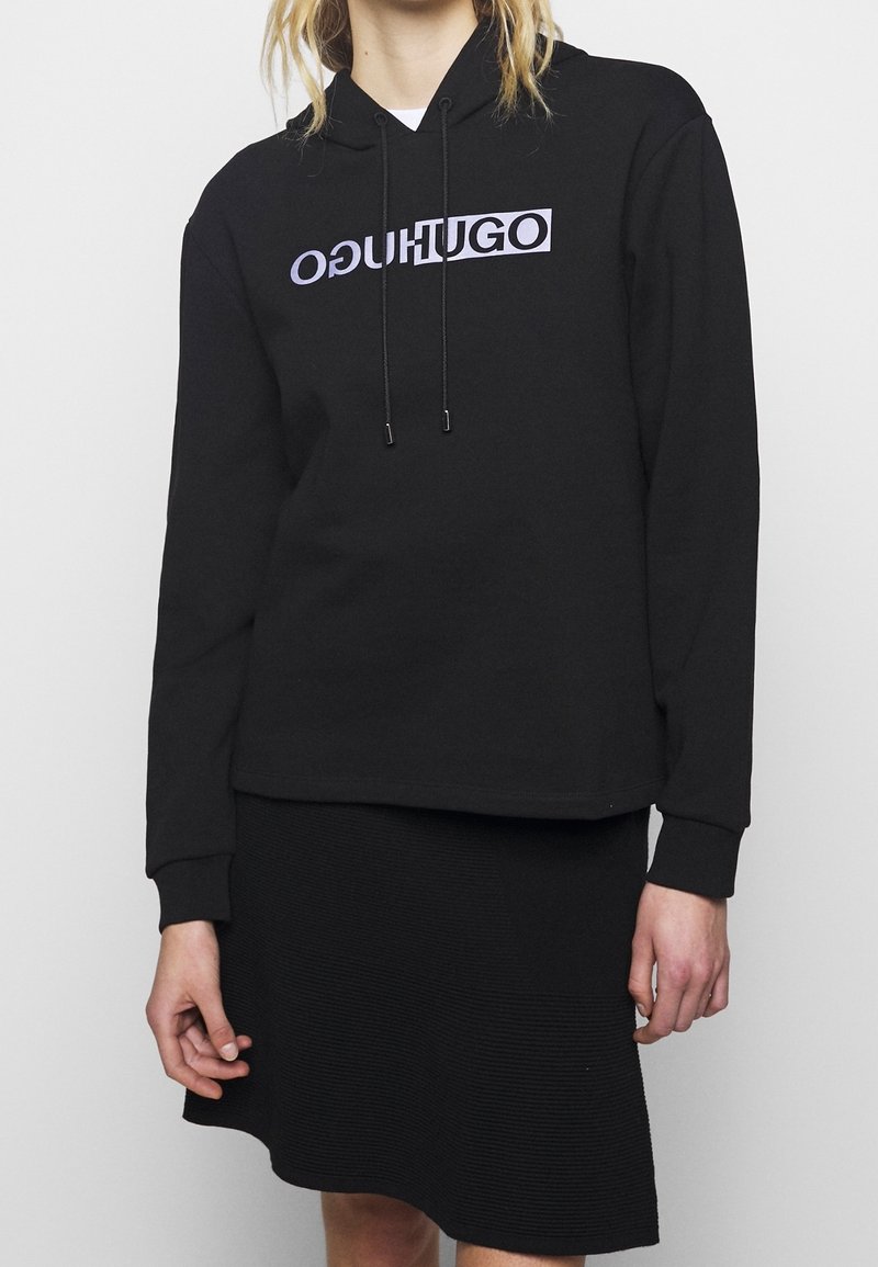 Personne portant un sweat à capuche noir avec le texte inversé "HUGO" en violet et une jupe noire texturée sur un fond clair uni.