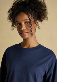 Navyblaue Langarm-T-Shirt mit rundem Halsausschnitt und lockerer Passform, aus glattem Stoff gefertigt. Das Modell hat lockige Haare und einen neutralen Hintergrund.