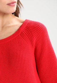 Red knitted sweater with ribbed texture, wide neckline, and long sleeves. The material appears soft and slightly stretchy.