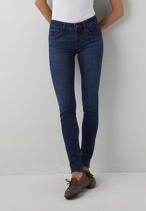 Femme portant un jean skinny bleu foncé, un haut blanc sans manches et des chaussures marron à lacets, debout contre un mur gris clair.