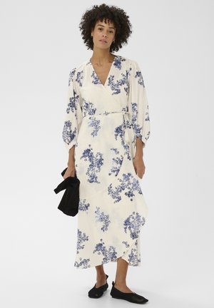 Kvinde iført en cremefarvet omslagskjole med blåt blomsterprint og trekvartlange ærmer, bærende en sort taske og sorte mesh-flade sko.