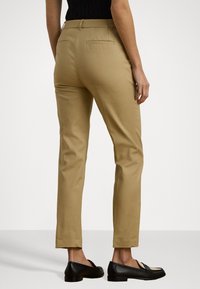 Beige-sävyiset räätälöidyt housut, joissa on slim fit -malli, litteä etuosa ja takataskut. Mukana mustat liukuhankalia. Sileä kankaan tekstuuri.