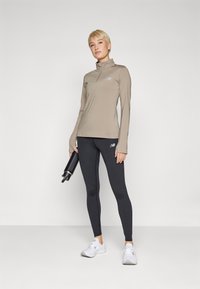 Haut de sport beige à manches longues avec un col haut et un logo, associé à un legging noir. Le mannequin tient une bouteille d'eau noire. Baskets blanches.
