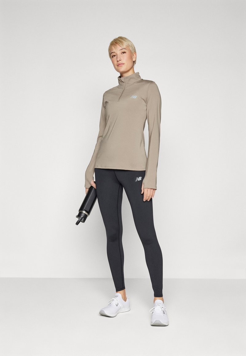 Beige Langarm-Sportoberteil mit hohem Kragen und Logo, kombiniert mit schwarzen Leggings. Das Model hält eine schwarze Wasserflasche. Weiße Sneakers.